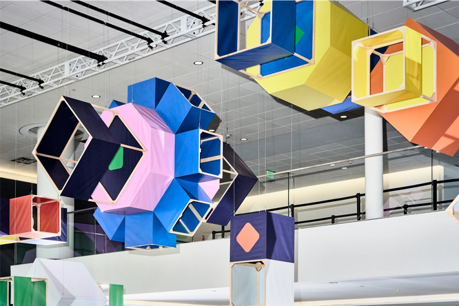 Six box kites are hanging from ceiling rafters over a large conference space. Each kite is a different 3D shape, with diamonds, squares, octagons, and one large asterisk. Some panels of the box kites are filled with blue, navy, yellow, or orange fabric, and some panels are empty.