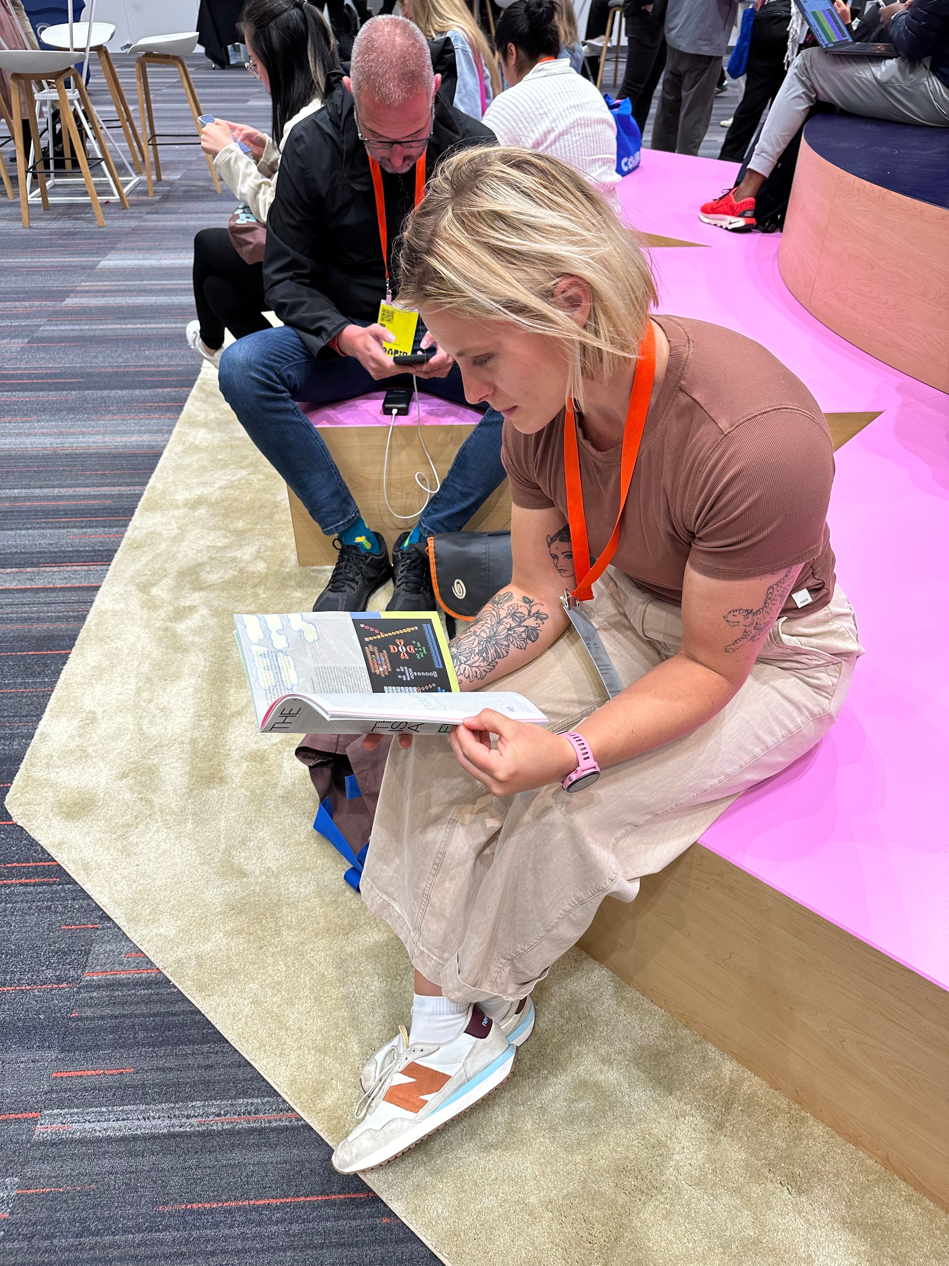 A blonde woman sits on a pink dias and reads The Prompt magazine.