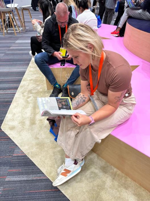 A blonde woman sits on a pink dias and reads The Prompt magazine.