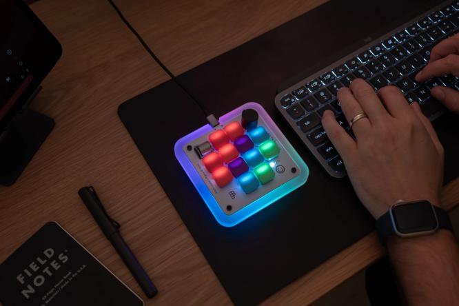 The Figma Creator Micro keyboard on a desk next to someone typing on a QWERTY keyboard.