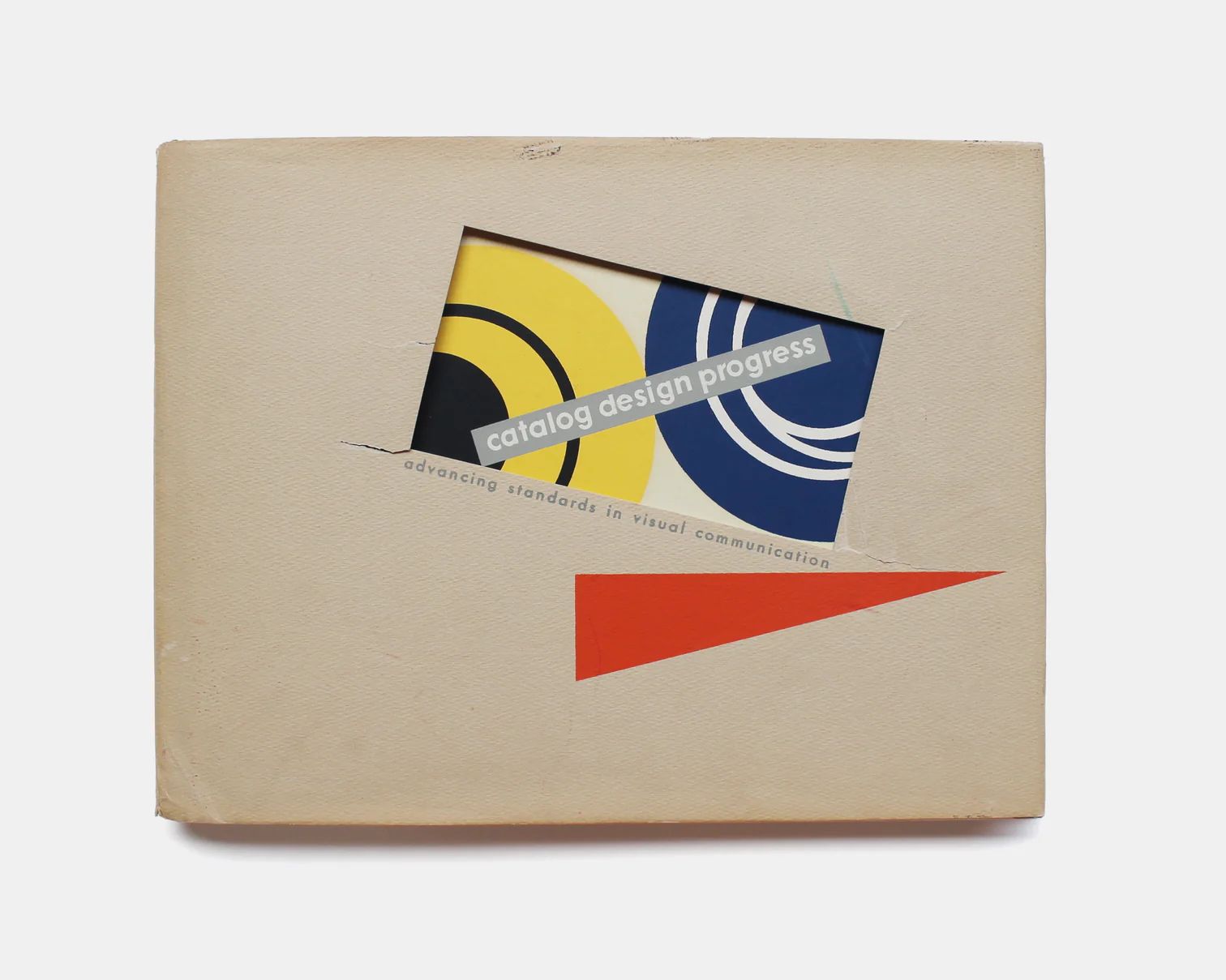 Vintage book cover with a die-cut window revealing a colorful abstract design underneath, featuring bold yellow, black, and blue geometric shapes. Text on the window reads “catalog design progress,” and smaller text below says “advancing standards in visual communication.” A red triangular shape is printed on the beige outer cover.