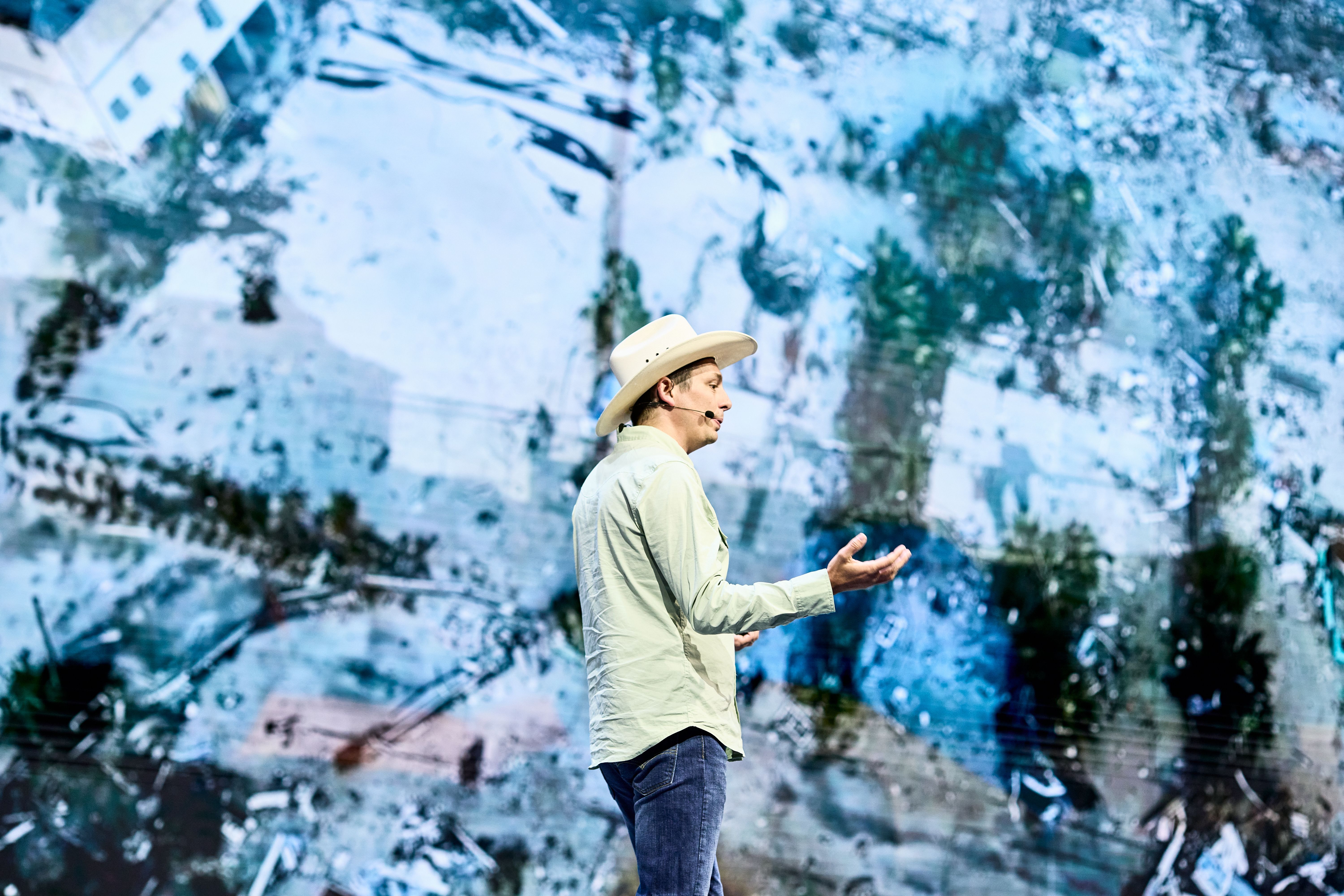A speaker in a light green shirt and cowboy hat gestures while presenting on stage. The background shows an aerial image of a landscape with apparent storm damage.