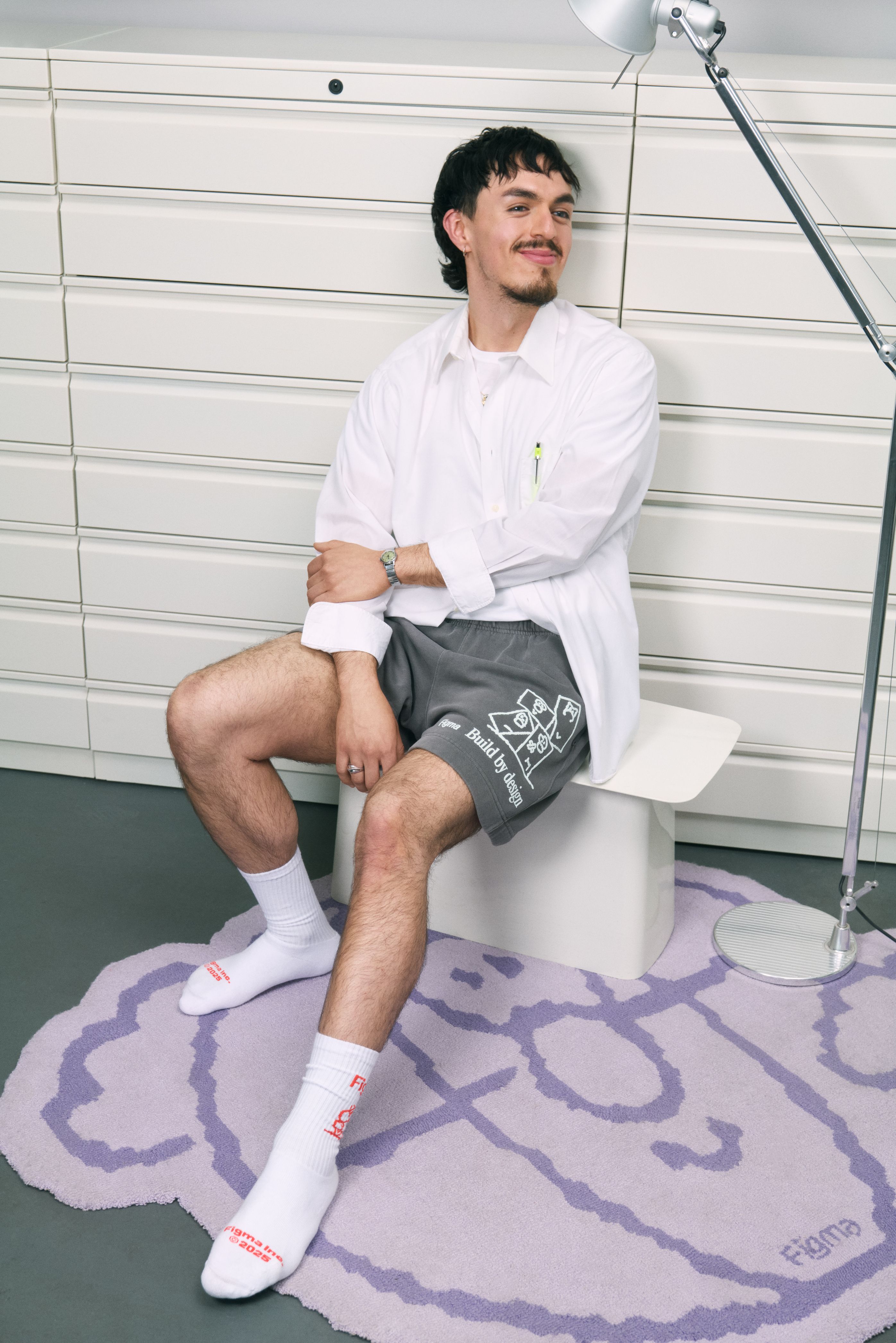 Man in white shirt and gray shorts sits smiling on stool beside lamp.