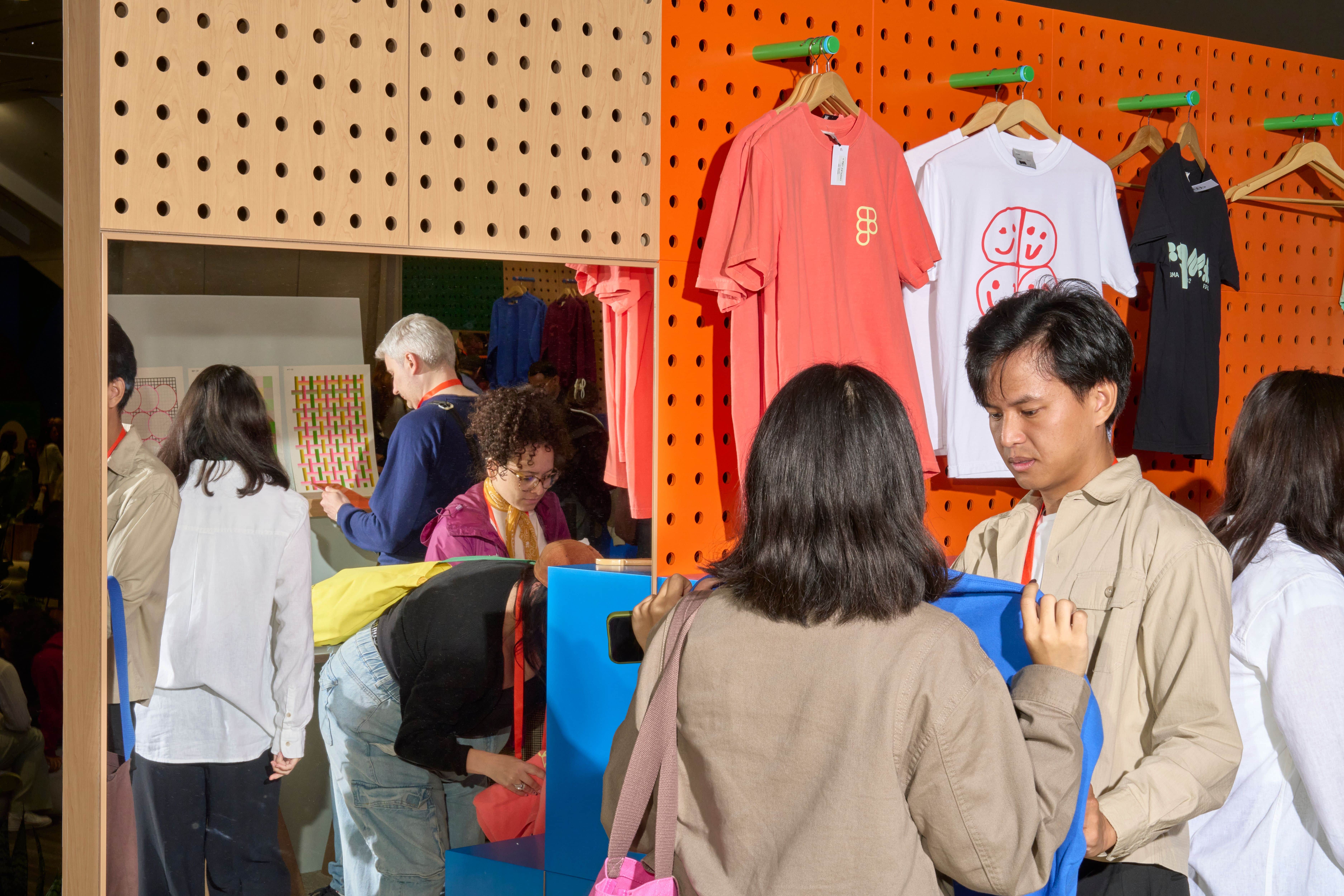 Shoppers hold up sweaters against  a backdrop of colorful tees hung on a pegboard wall.