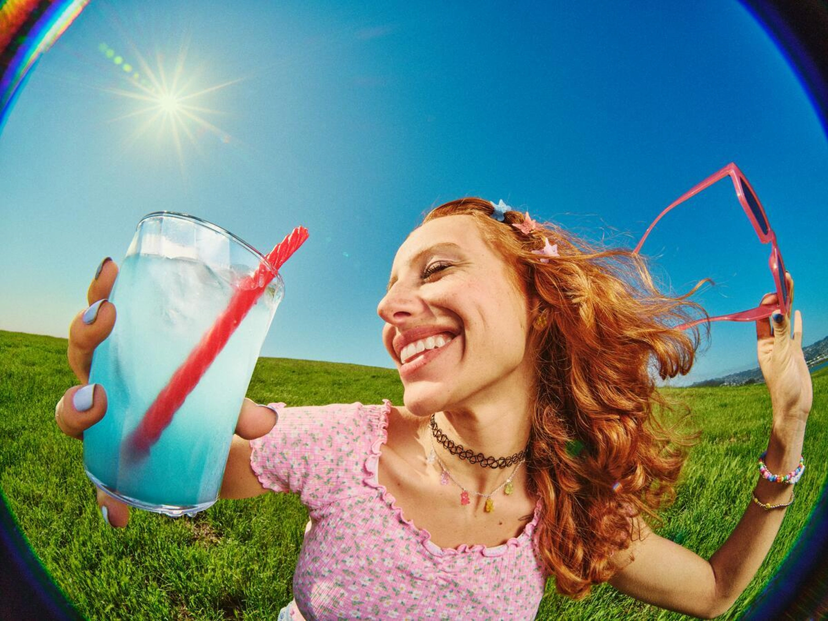 Fish-eye photo of a smiling person with butterfly clips in their hair enjoying a sunny day outdoors, holding a bright turquoise drink with a licorice candy used as a straw.