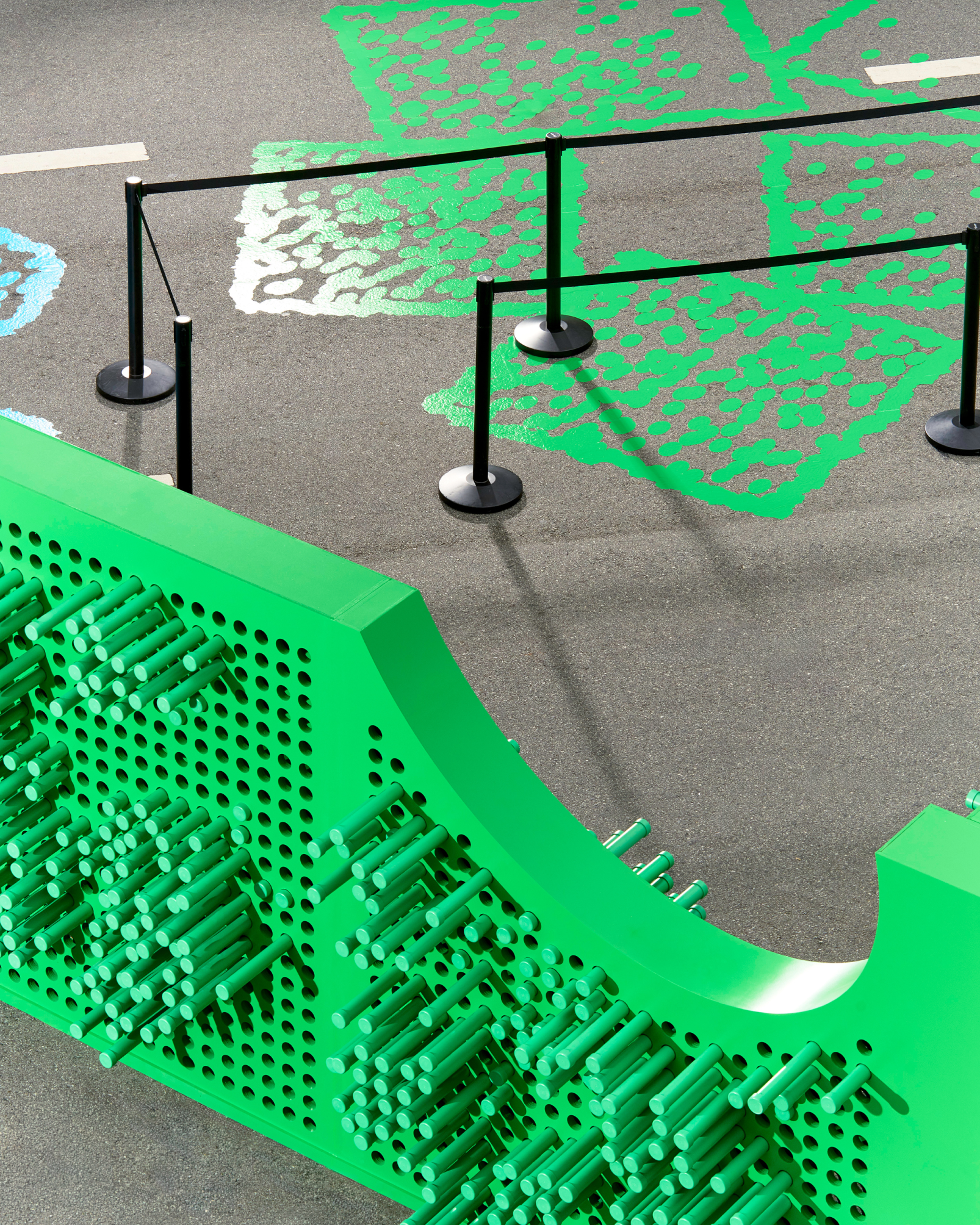 A green pin-art installation made of protruding cylindrical pegs sits on pavement, cordoned off by black stanchions, with a scattered green dot pattern painted on the ground behind it.