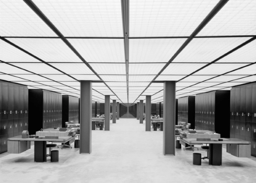 A vast, symmetrical office filled with rows of desks and filing cabinets stretches into the distance under a glowing grid ceiling.