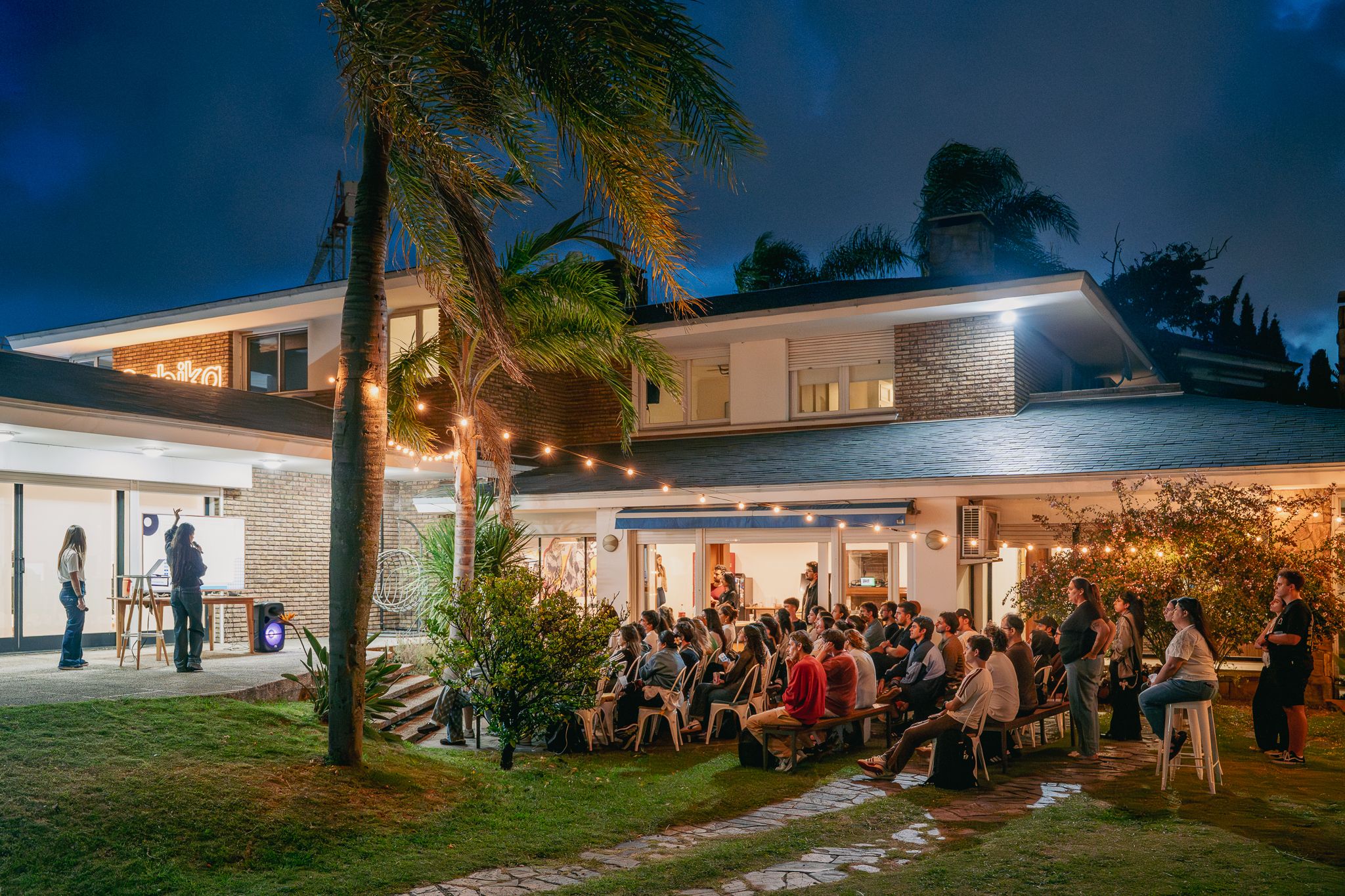 Attendees sit in front of a screen at an outdoor venue featuring an evening sky and palm trees