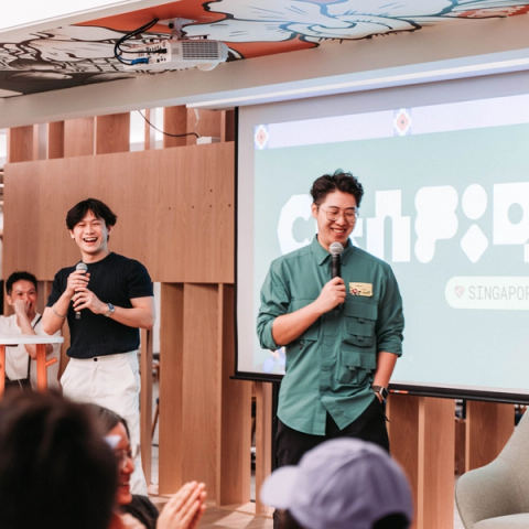 Two speakers holding microphones stand on a stage in front of a screen with the word "Config" and "Singapore" displayed. They are smiling and addressing an audience in a modern indoor setting.
