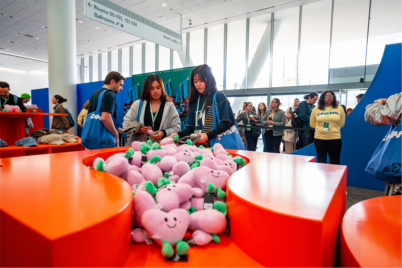 Event attendees browse orange tables filled with smiling pink heart-shaped plush toys, while others wait in line under a bright convention lobby sign.