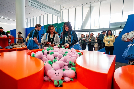 Event attendees browse orange tables filled with smiling pink heart-shaped plush toys, while others wait in line under a bright convention lobby sign.