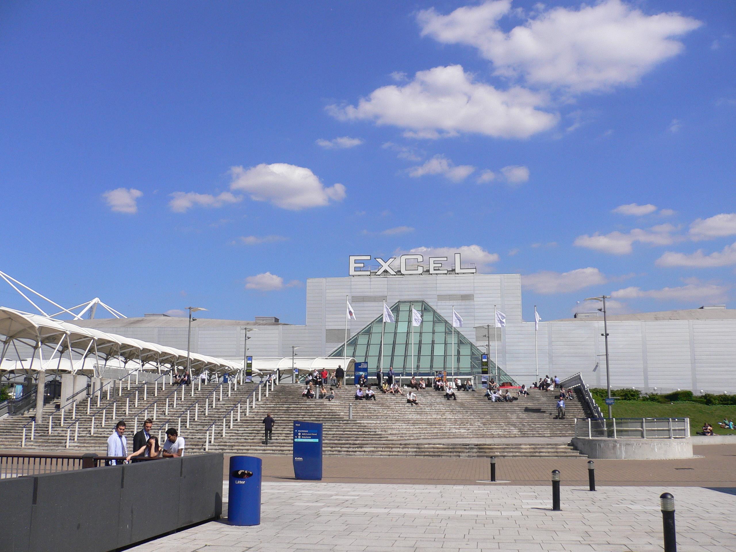 The exterior of the ExCeL Center, featuring a large glass entrance, white canopies, and steps with people sitting or walking under a sunny blue sky.