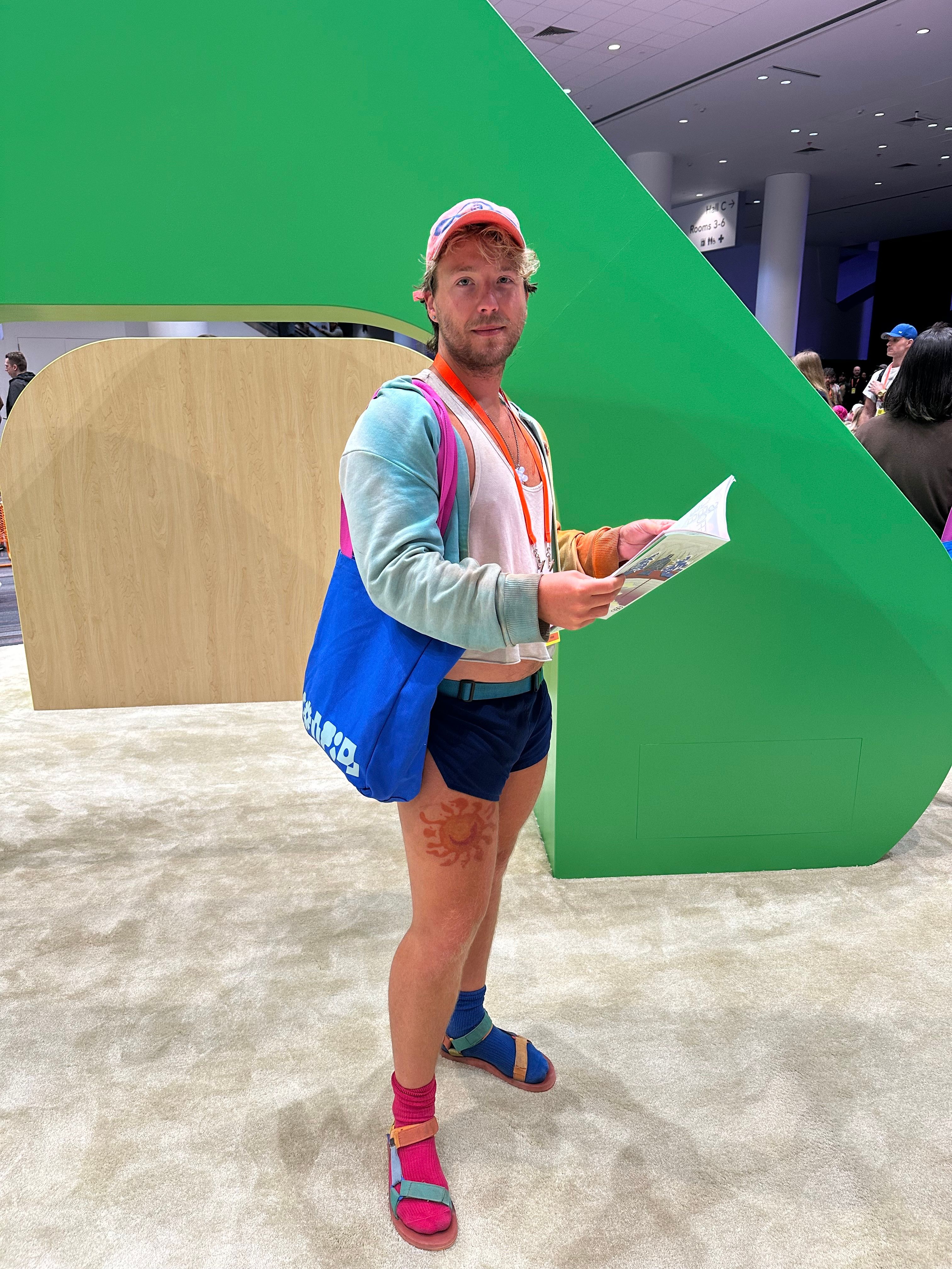 A man wears a white crop tee, a blue cardigan, blue shorts, and colorful mismatched socks