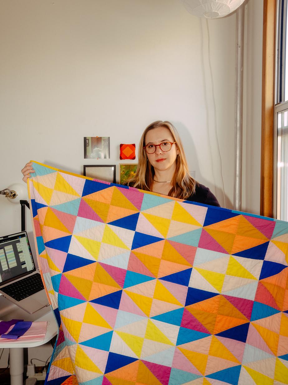 Nicole, who has blonde hair and wears red glasses, holds up a quilt made up of teal, orange, yellow, and blue triangles.