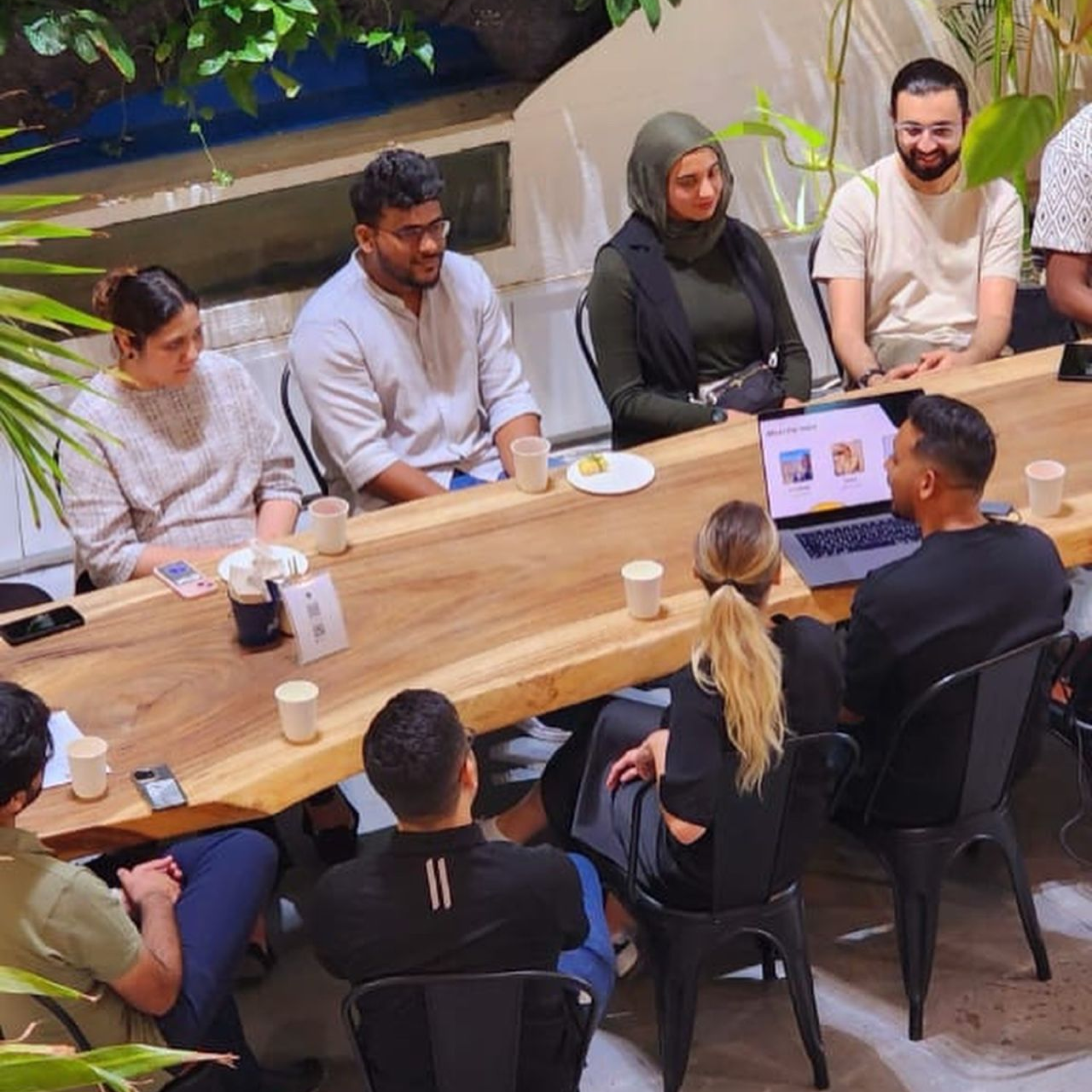 A group of people seated around a long wooden table at a gathering. They appear to be engaged in conversation, with laptops, cups, and plates on the table. The setting is surrounded by plants, giving a cozy, casual atmosphere.