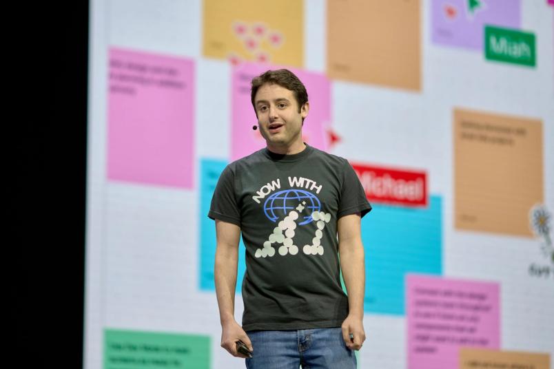Dylan Field stands in front of a screen wearing a shirt that reads, “Now with AI.”