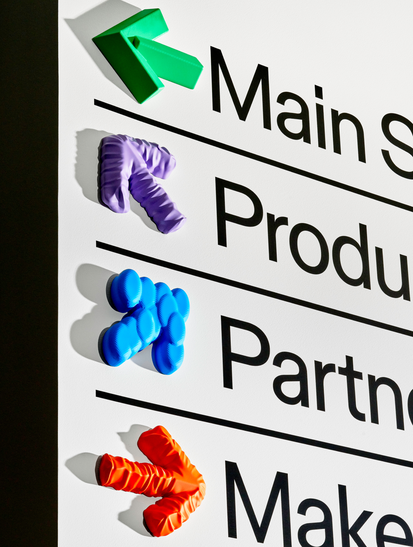 A white wayfinding sign lists directions labeled Main S..., Produ..., Partn..., and Make..., each marked with a colorful sculptural 3D arrow in green, purple, blue, and orange.