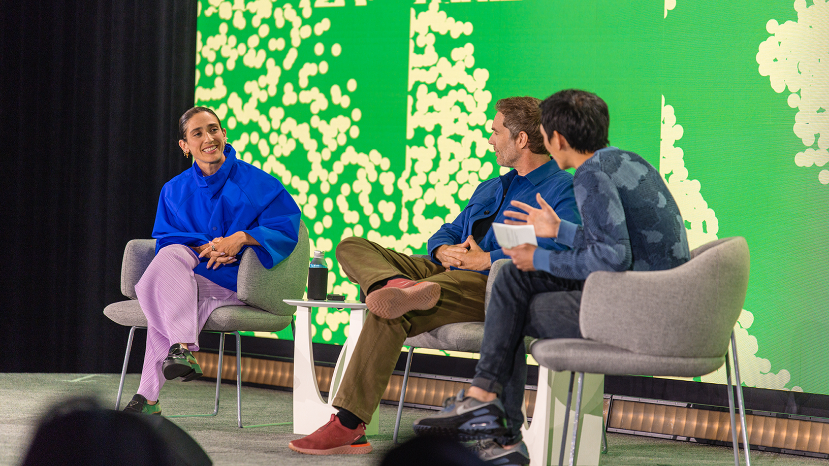 Rachel Been, Charlie Sutton, and Yuhki Yamashita sit in gray armchairs on a stage in front of a bright green patterned screen, mid-conversation.