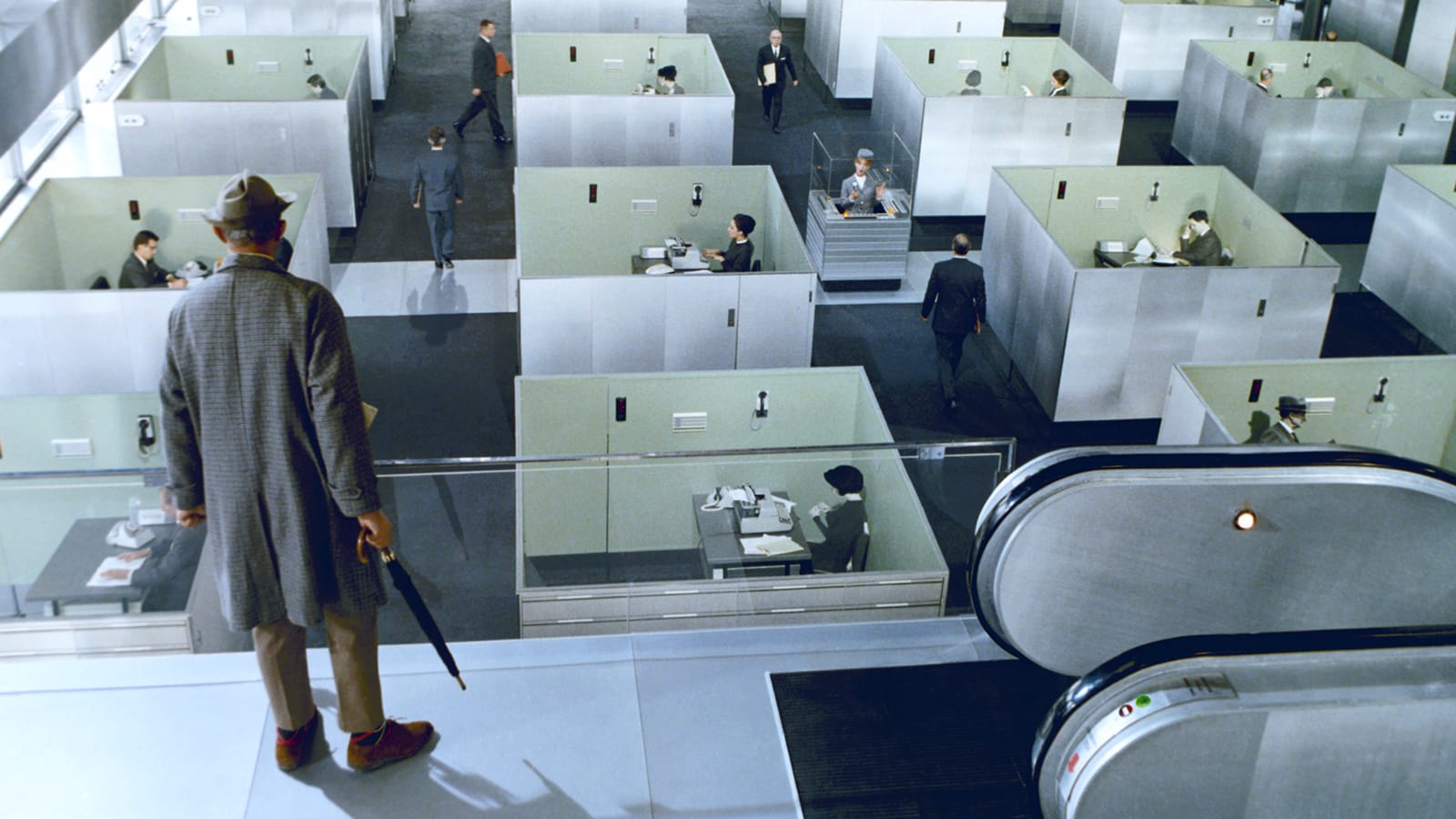 A man in a coat and hat stands at the top of an escalator, overlooking a grid of identical gray office cubicles filled with workers.