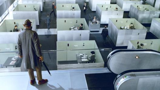 A man in a coat and hat stands at the top of an escalator, overlooking a grid of identical gray office cubicles filled with workers.