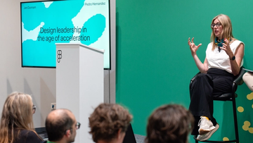 A speaker gestures from a stool beside a slide titled "Design leadership in the age of acceleration," addressing a seated audience.