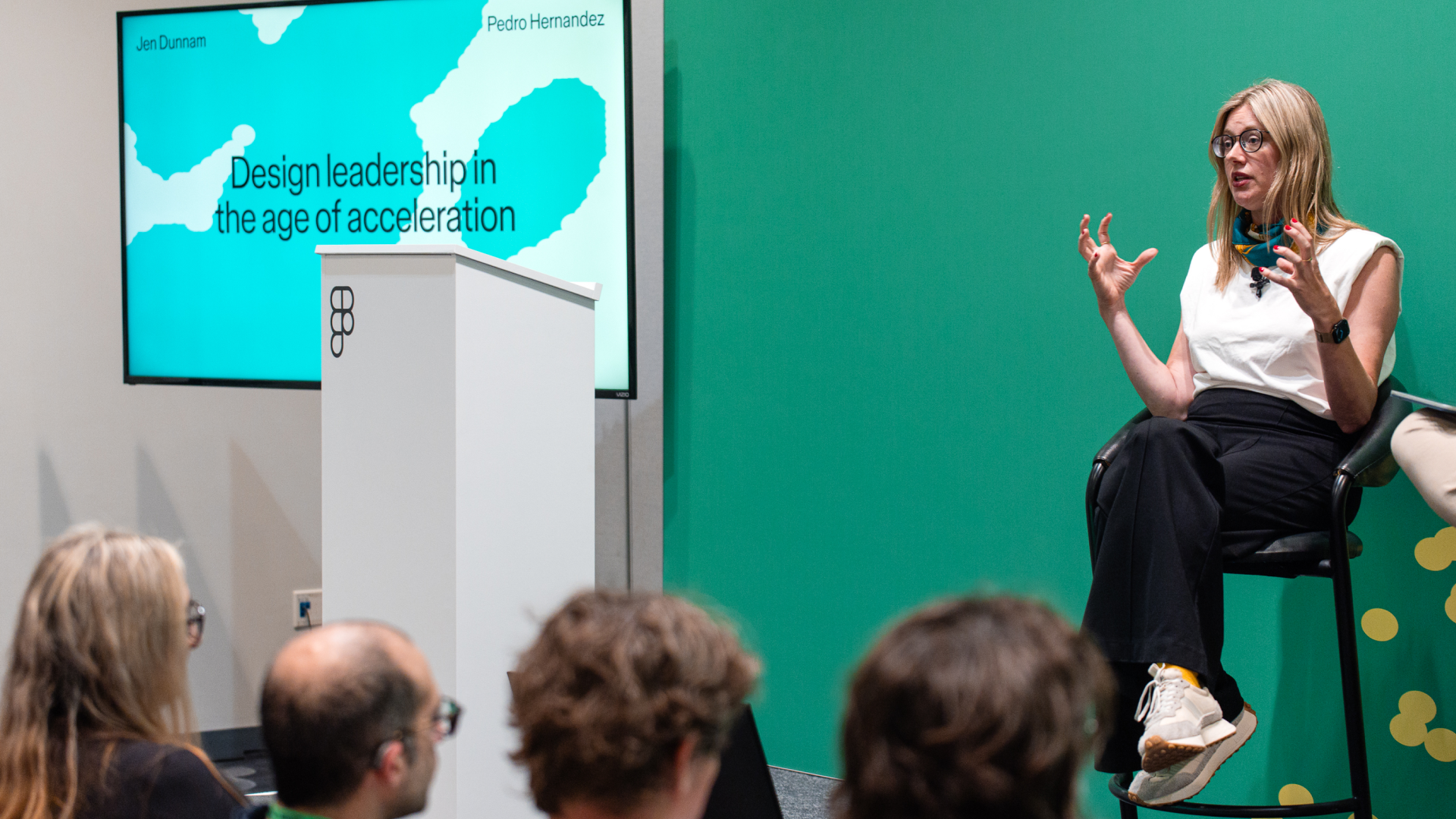 A speaker gestures from a stool beside a slide titled "Design leadership in the age of acceleration," addressing a seated audience.