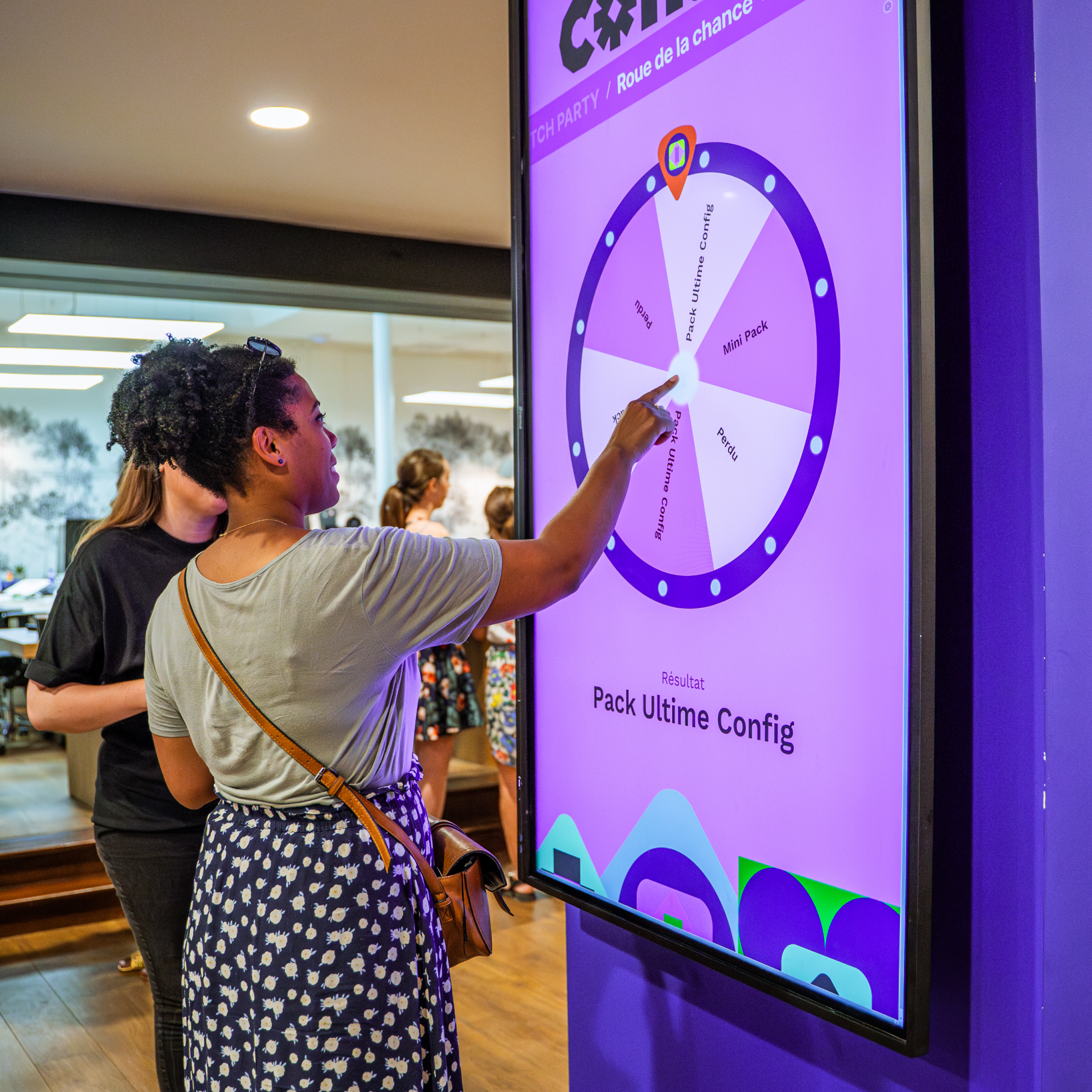 A woman points at a digital wheel of fortune displayed on a large vertical screen. The screen has a purple background, with sections of the wheel labeled in French and event-related terms. Other attendees can be seen in the background.