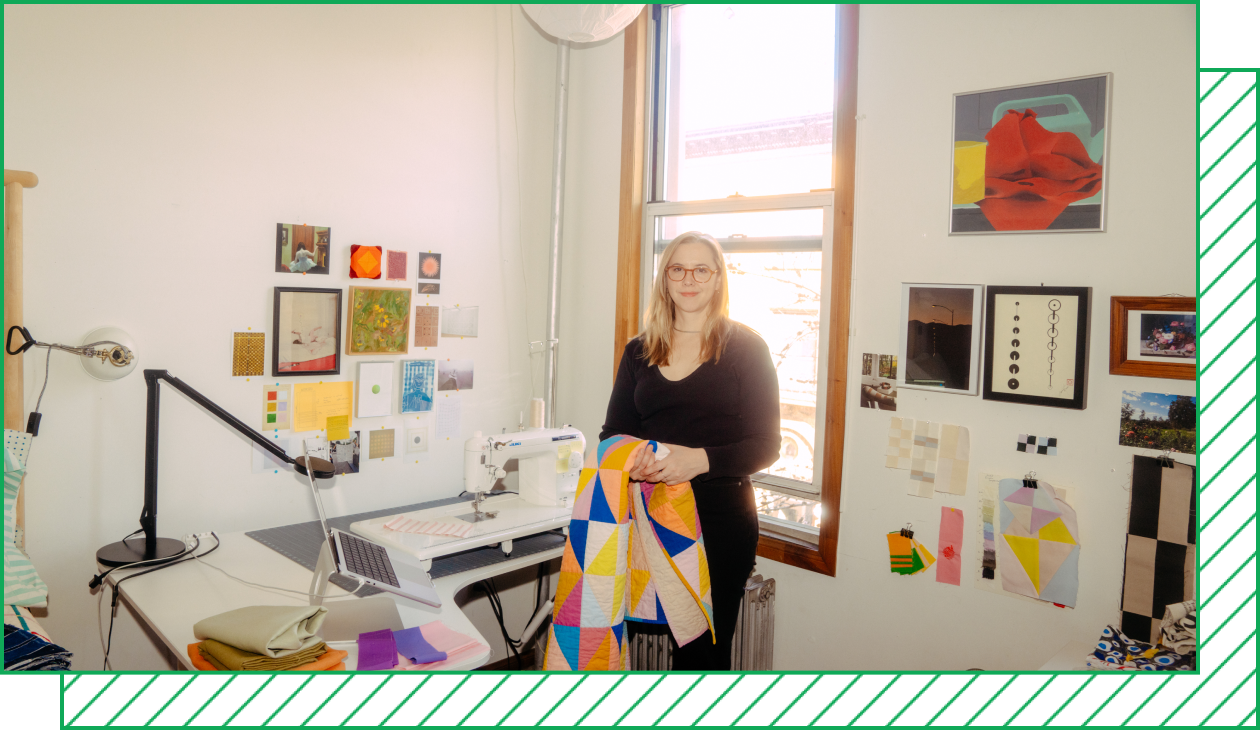 A photograph shows Nicole Boettcher in her bedroom studio, which has a desk with a sewing machine and fabric swatches and art pinned to the wall. She stands in front of a window with a quilt folded over her arm.