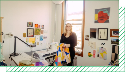 A photograph shows Nicole Boettcher in her bedroom studio, which has a desk with a sewing machine and fabric swatches and art pinned to the wall. She stands in front of a window with a quilt folded over her arm.