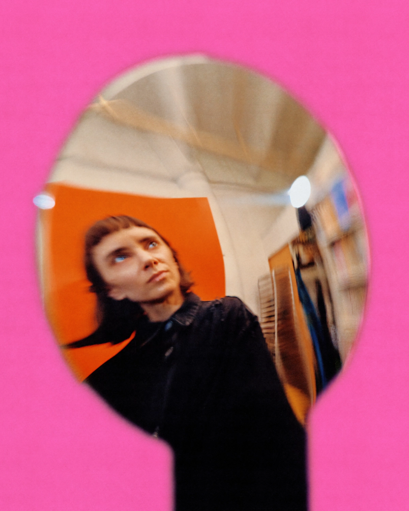 Distorted reflection of Kelsey in a shiny spoon shape against a bright pink background in studio.