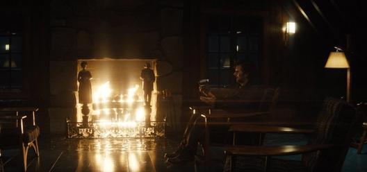A man sits alone by a roaring fire in a dark wood cabin, lit warmly by the flames and a single lamp.