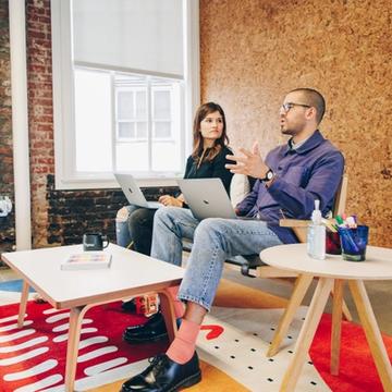 Two employees in an office room with their laptops talking to each other