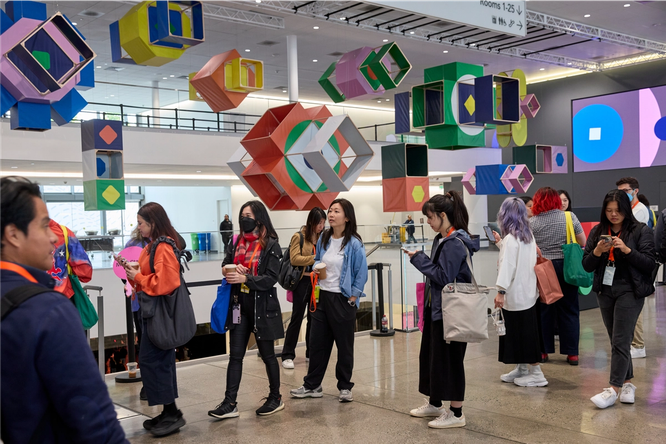 In the lobby of a large event space, there are people walking, talking to other people, and looking at their phones. Behind them, there are 14 hanging box kites, each differently sized, shaped, and colored.