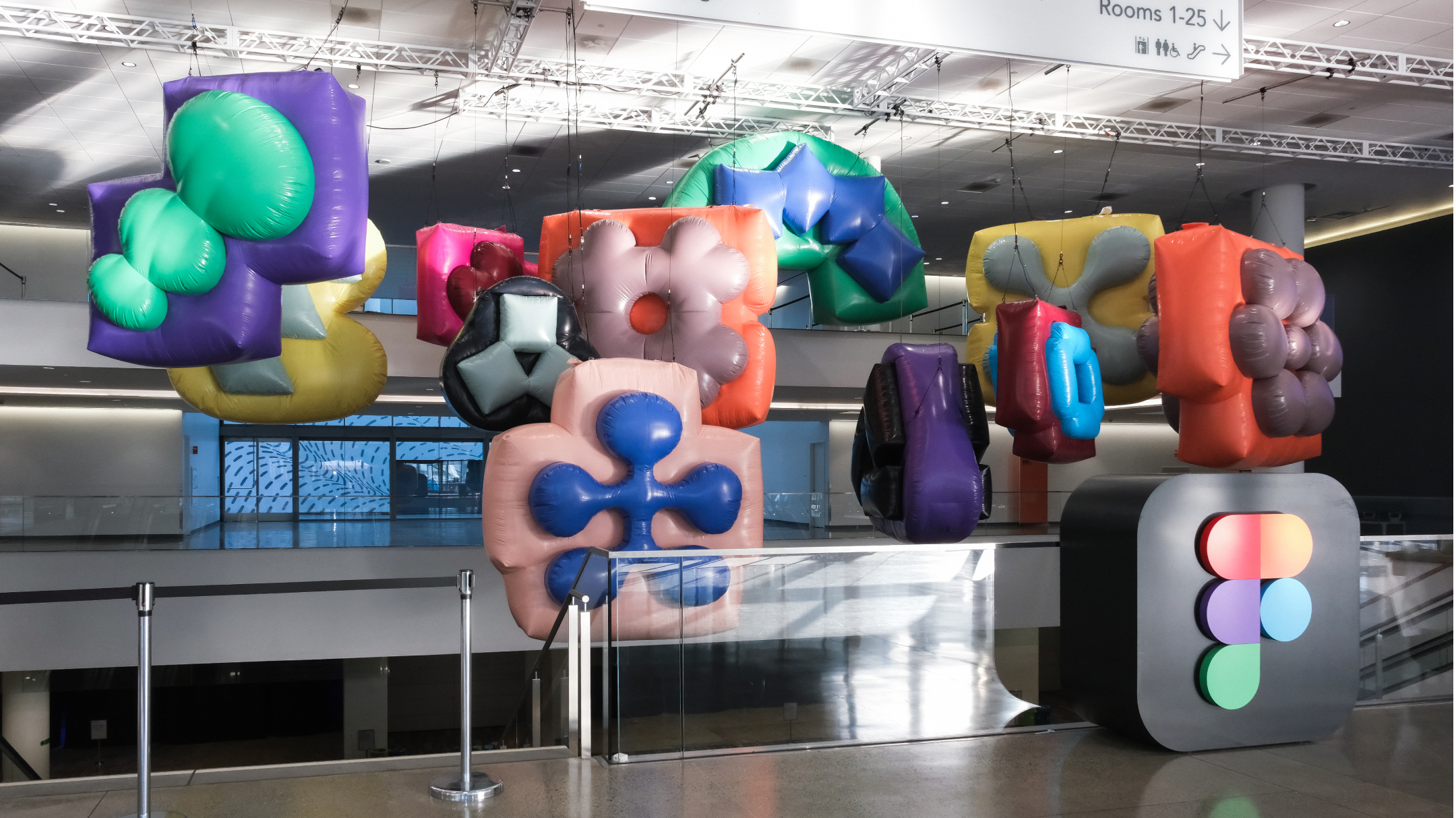 Colorful inflatable sculptures hang above a Figma logo pedestal inside a modern convention center space.