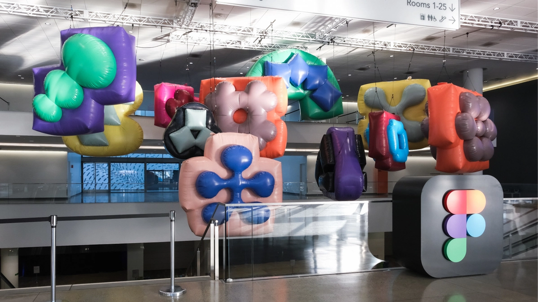 Colorful inflatable sculptures hang above a Figma logo pedestal inside a modern convention center space.