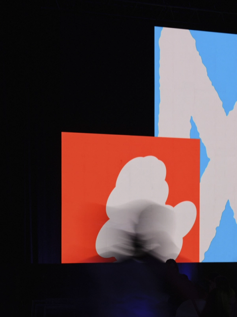 An on-stage screen shows abstract graphics with a blurred figure in front