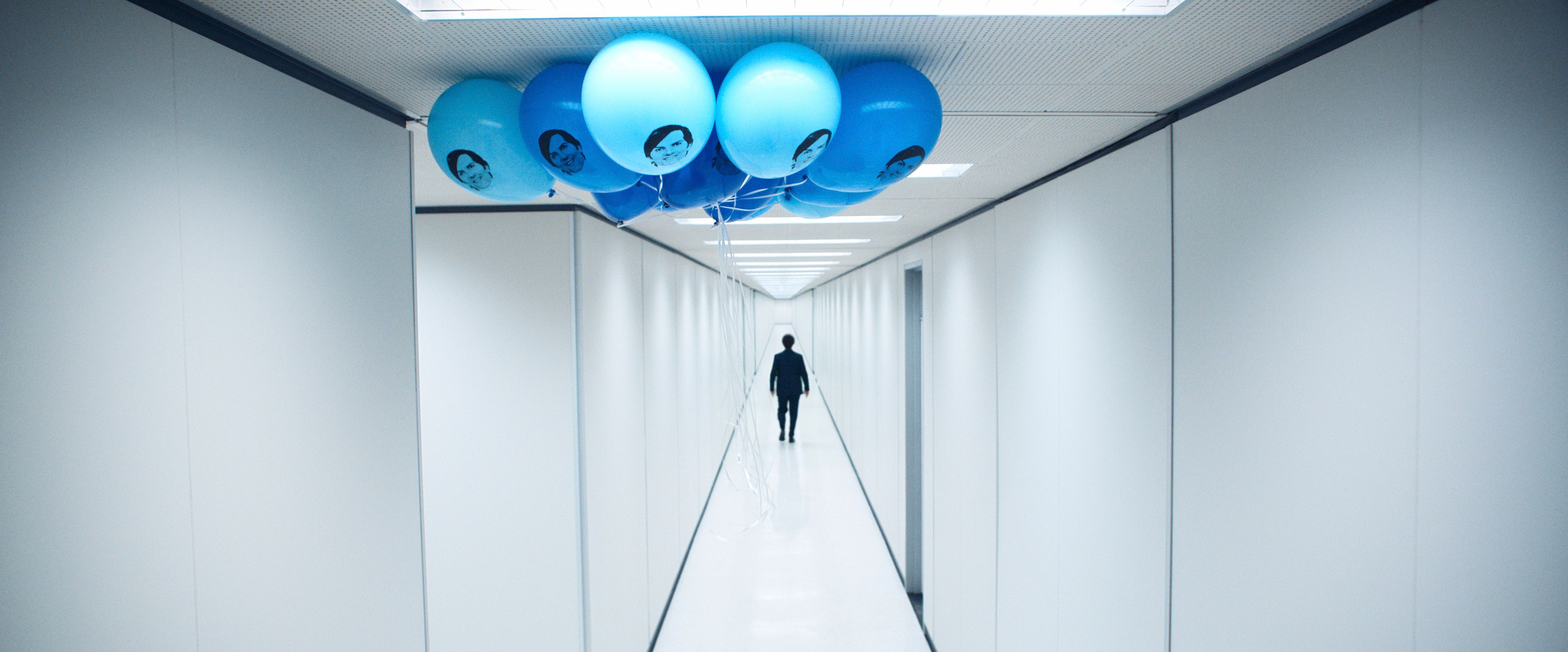 A person walks down a stark white hallway decorated with a cluster of blue balloons featuring a printed face on them.