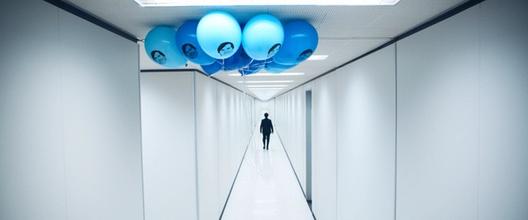 A person walks down a stark white hallway decorated with a cluster of blue balloons featuring a printed face on them.