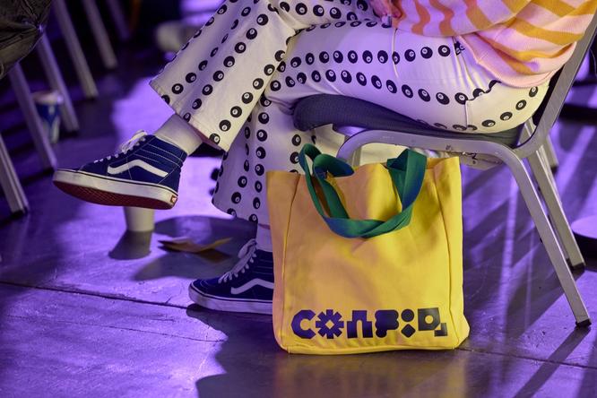 A yellow Config tote bag with green straps sits beside a conference attendee’s chair. The ambient purple lighting suggests they’re seated in the audience of an event talk.