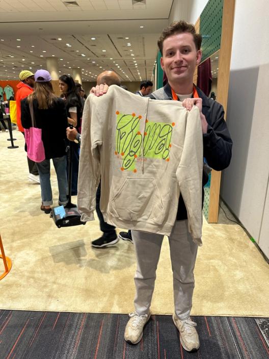 A man holds up a tan hoodie that says Figma in lime green bubble letters.