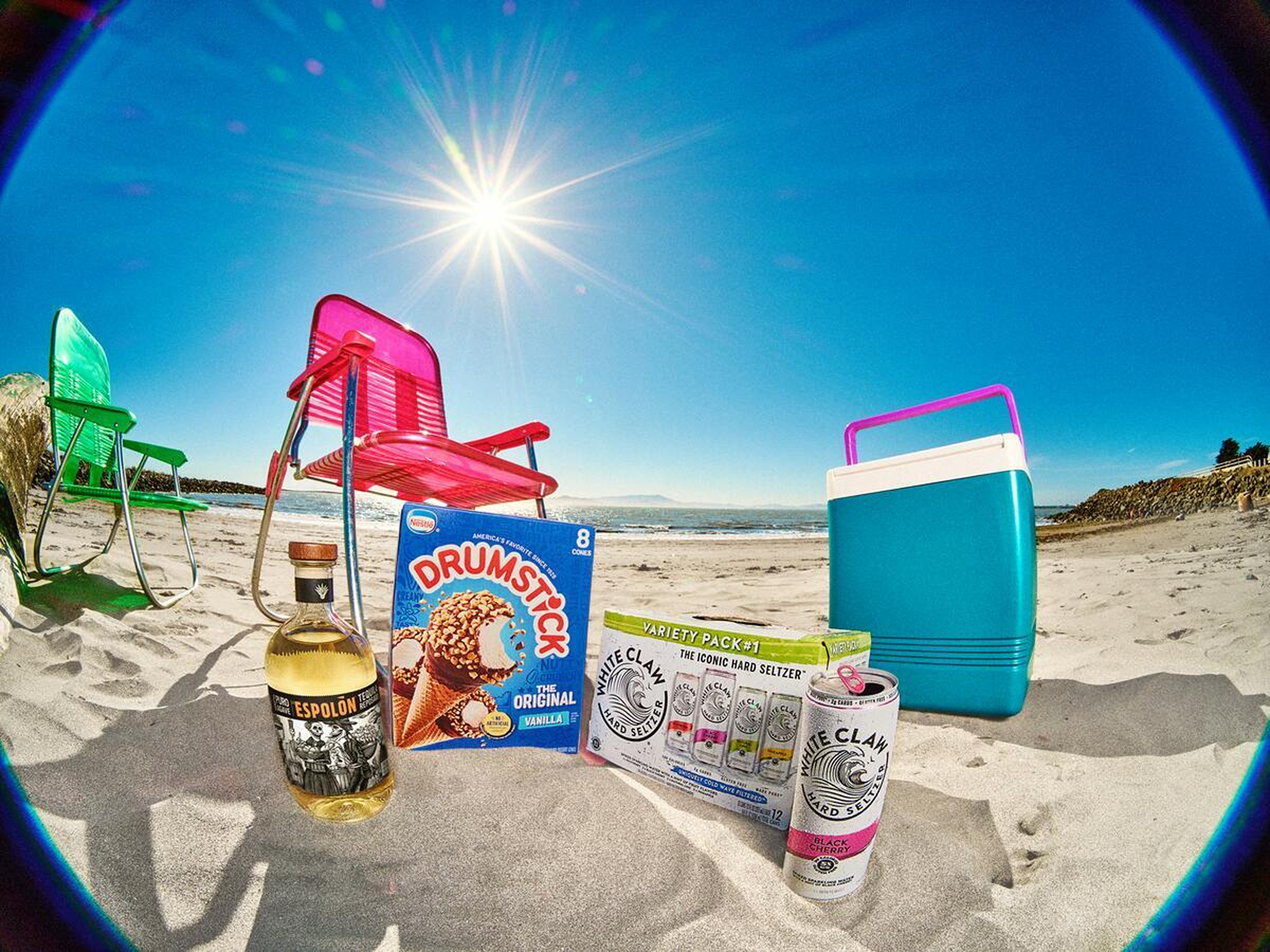 Colorful chairs, cooler, tequila, Drumstick cones, and White Claw seltzer arranged on sandy beach under sun.