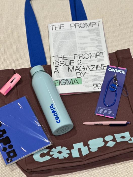 A swag flatlay shows a brown Config tote bag, a blue notepad, a blue water bottle, a charm keychain, pens, and a magazine.