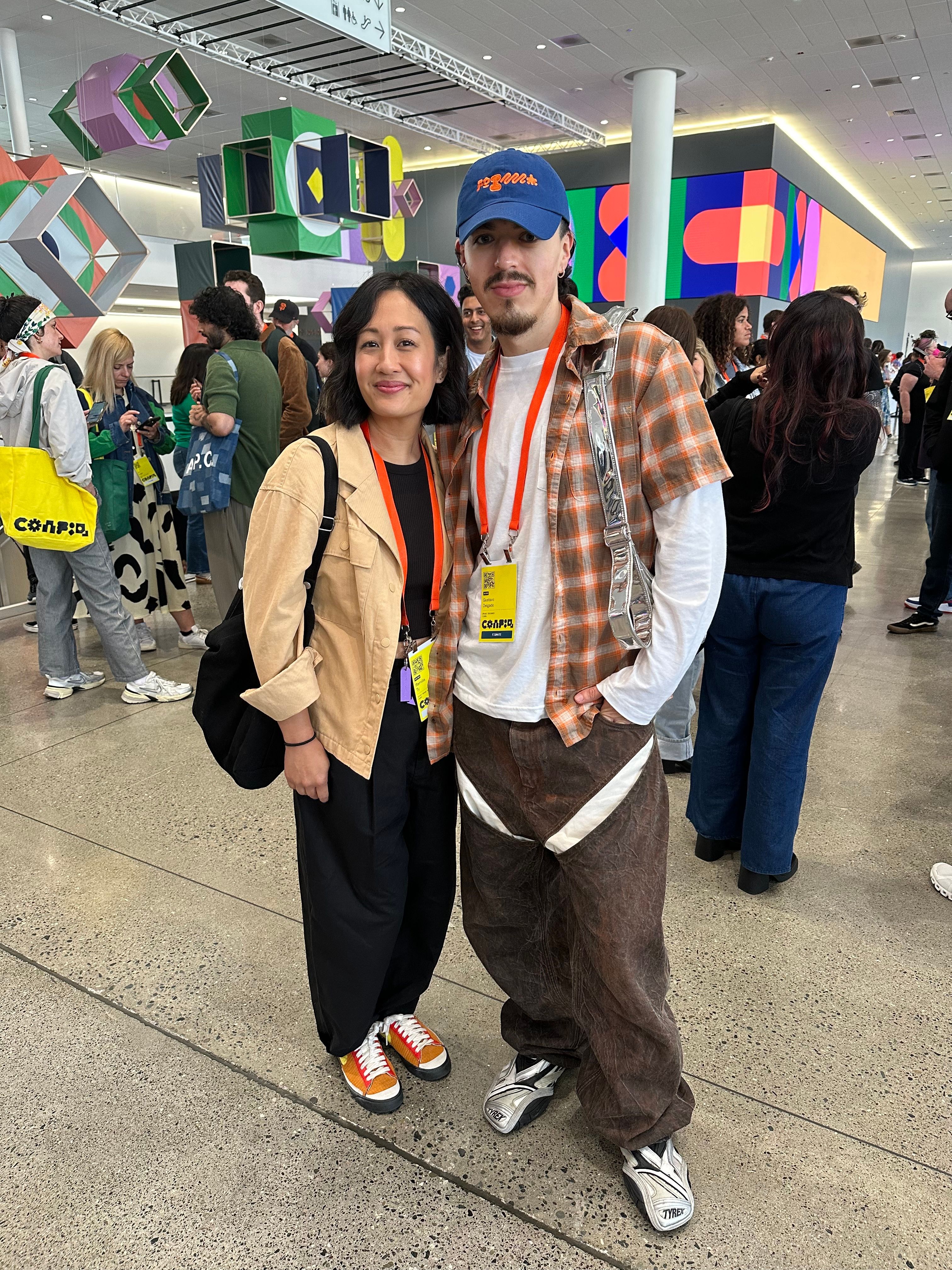 A woman wears a tan blazer and black pants, and a man wears a checkered button-down over a long sleeve white tee and a silver shoulder bag