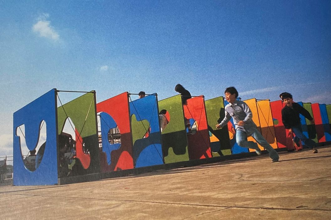 Children running and playing around a colorful, maze-like playground made up of large, flat panels. Each panel is painted in bold colors such as blue, red, green, and yellow, with abstract cutout shapes. The kids are energetically running in the foreground under a bright blue sky.