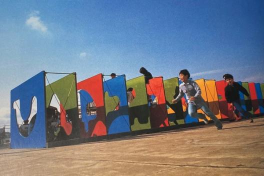 Children running and playing around a colorful, maze-like playground made up of large, flat panels. Each panel is painted in bold colors such as blue, red, green, and yellow, with abstract cutout shapes. The kids are energetically running in the foreground under a bright blue sky.