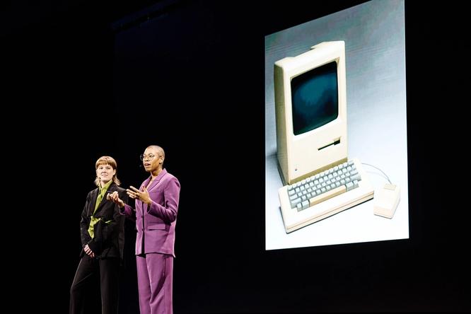 Two speakers stand side by side on stage. One is wearing a purple suit and gesturing while speaking. The other is dressed in a black and green outfit, looking at the audience. A large projected image of a vintage Macintosh computer is visible behind them.