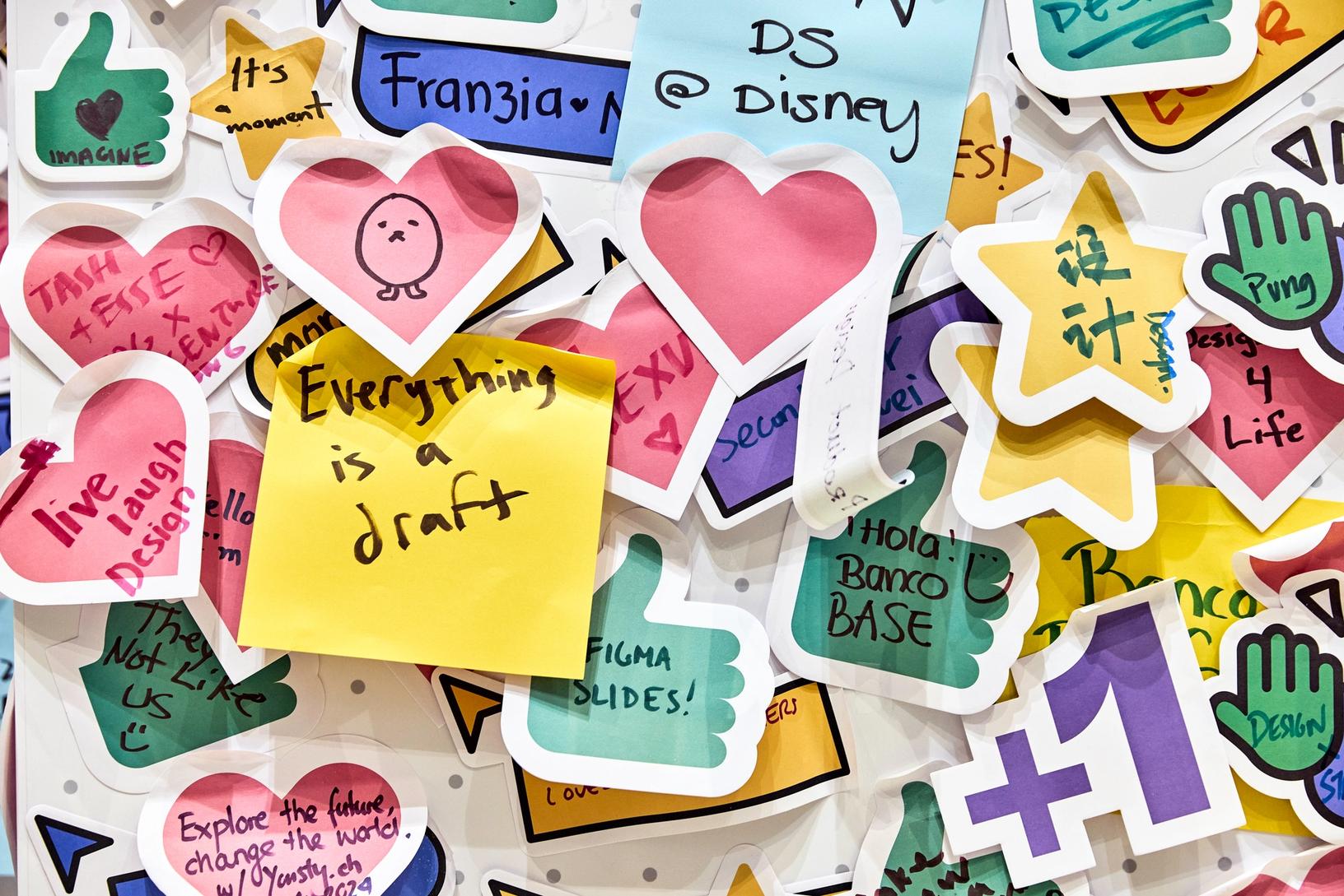 A colorful collage of various sticky notes and cut-out shapes with handwritten messages and doodles. The notes include hearts, stars, thumbs-ups, and speech bubbles. Prominent messages read "Everything is a draft" on a yellow note, "live laugh design" on a pink heart, and "Figma slides!" on a green thumbs-up. The collection exudes creativity and positivity, with phrases and symbols written in different colors.