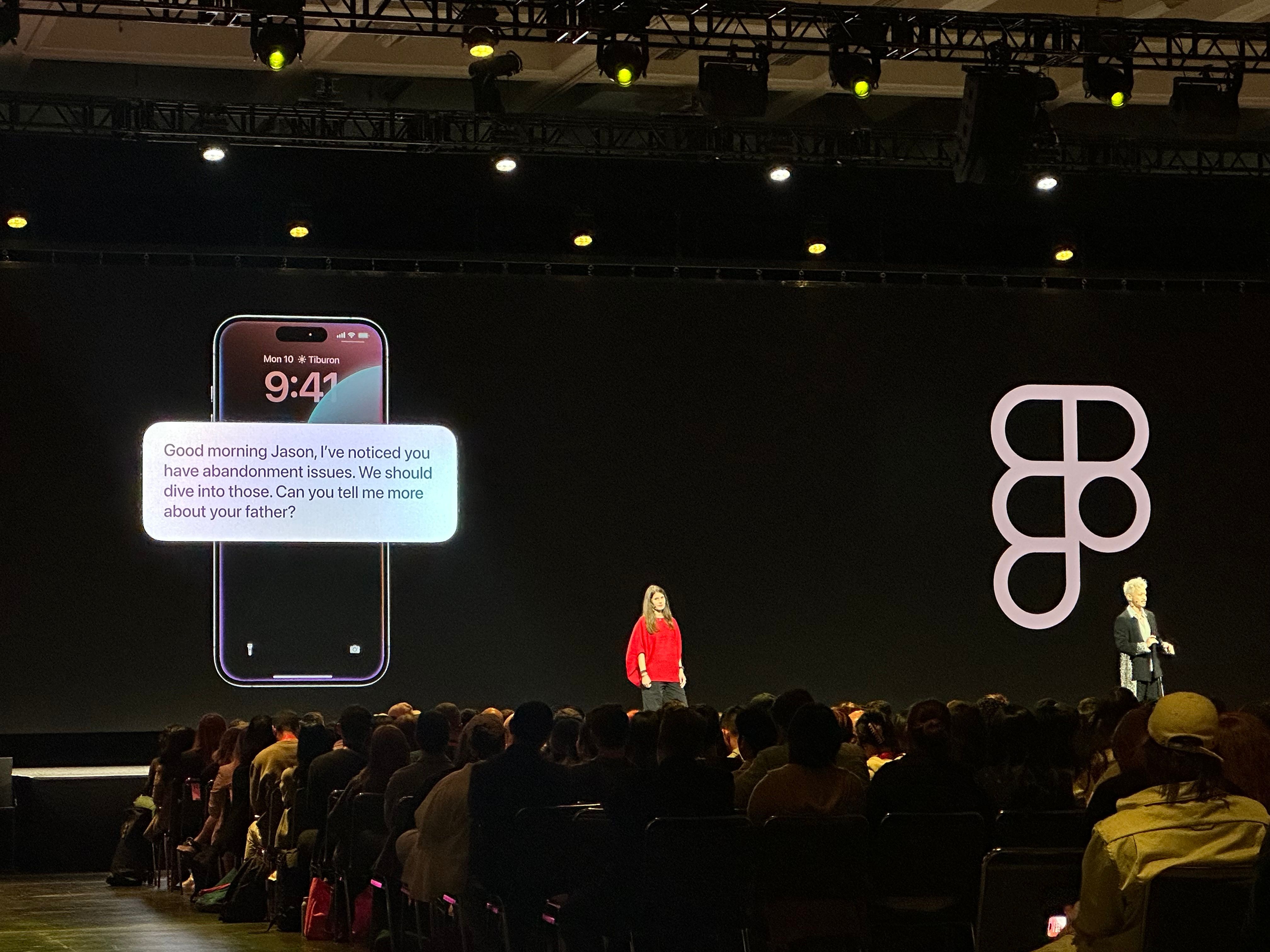 Two people are on stage. On the screen, a message on a phone reads: “Good morning, Jason, I’ve noticed you have abandonment issues. We should dive into those. Can you tell me more about your father?”