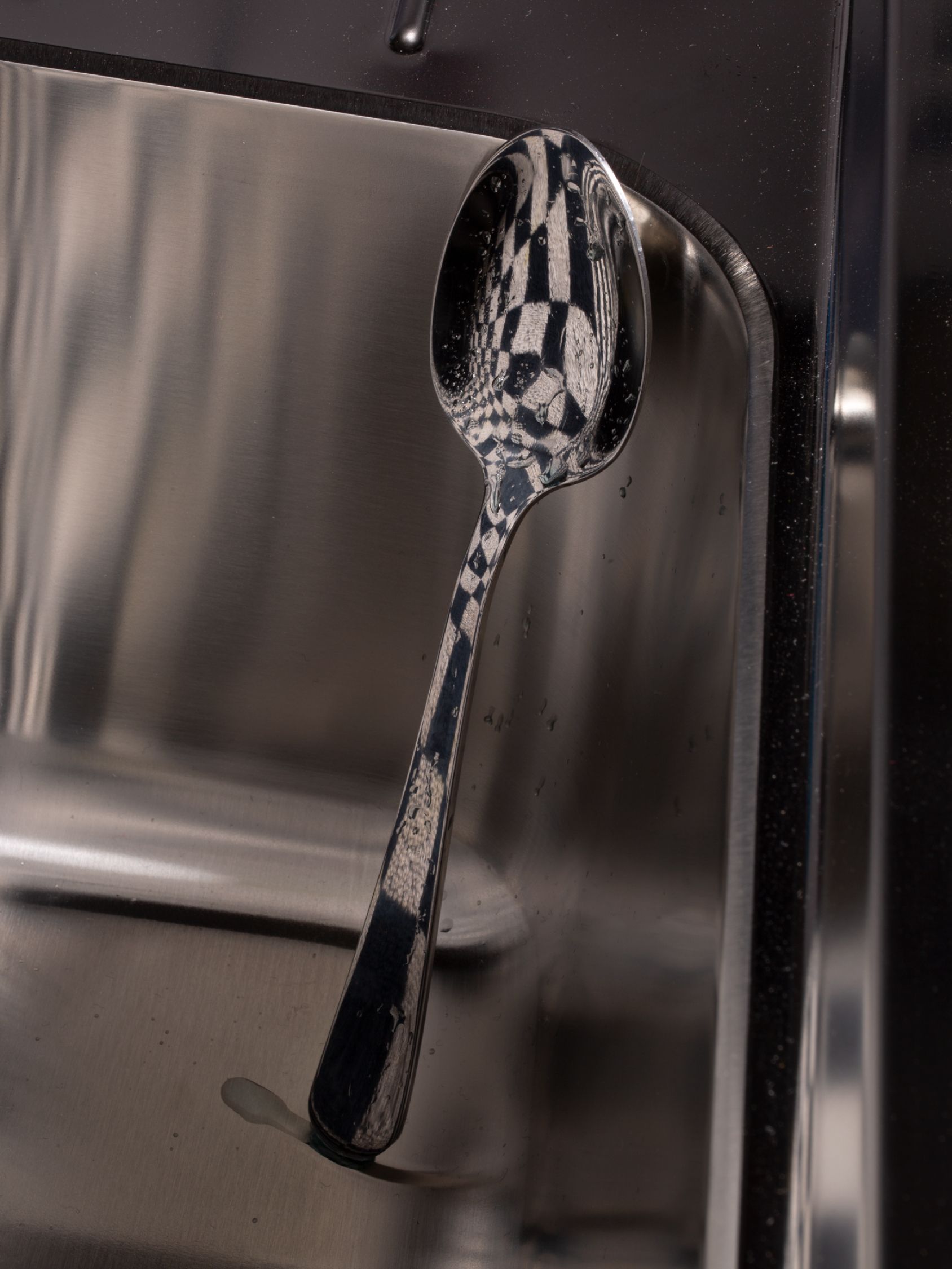 Reflective metal spoon balanced inside a stainless-steel sink, mirroring a distorted checkerboard pattern in its bowl.