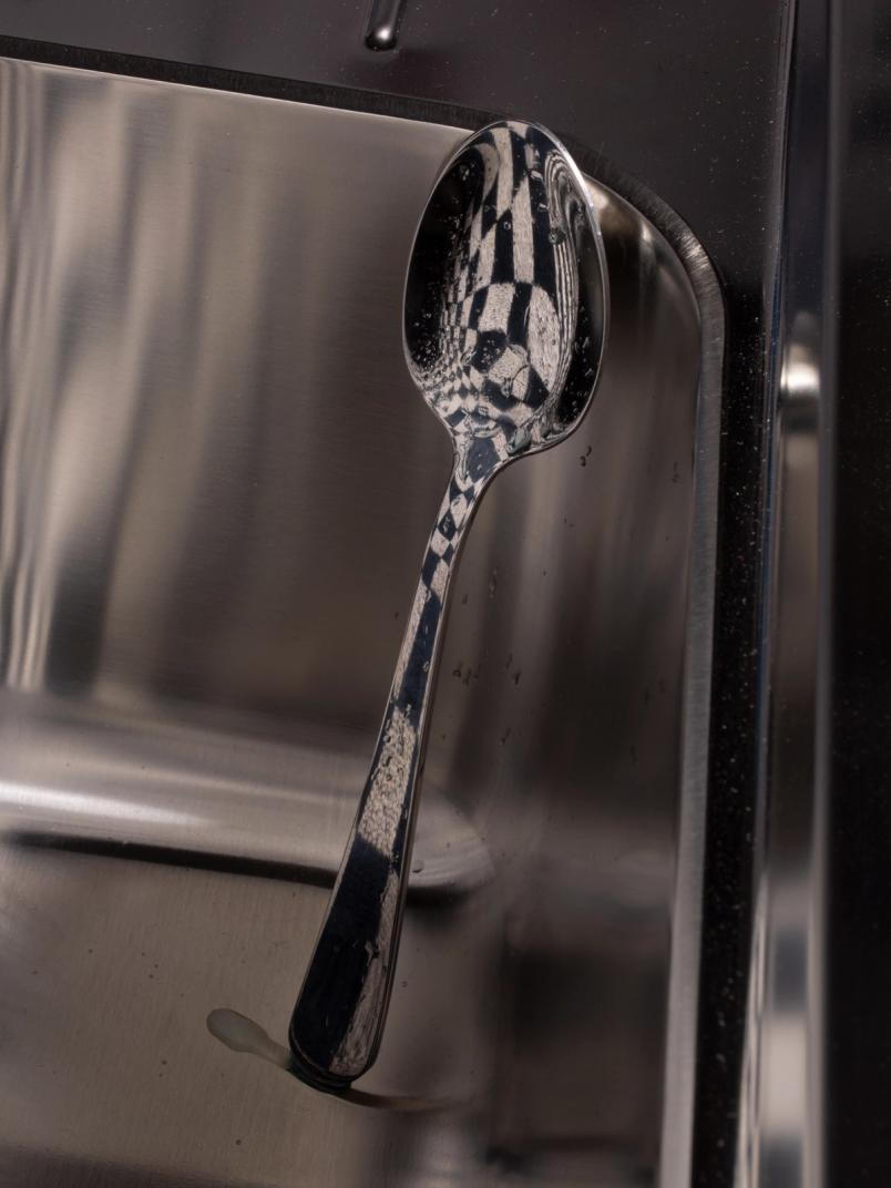 Reflective metal spoon balanced inside a stainless-steel sink, mirroring a distorted checkerboard pattern in its bowl.